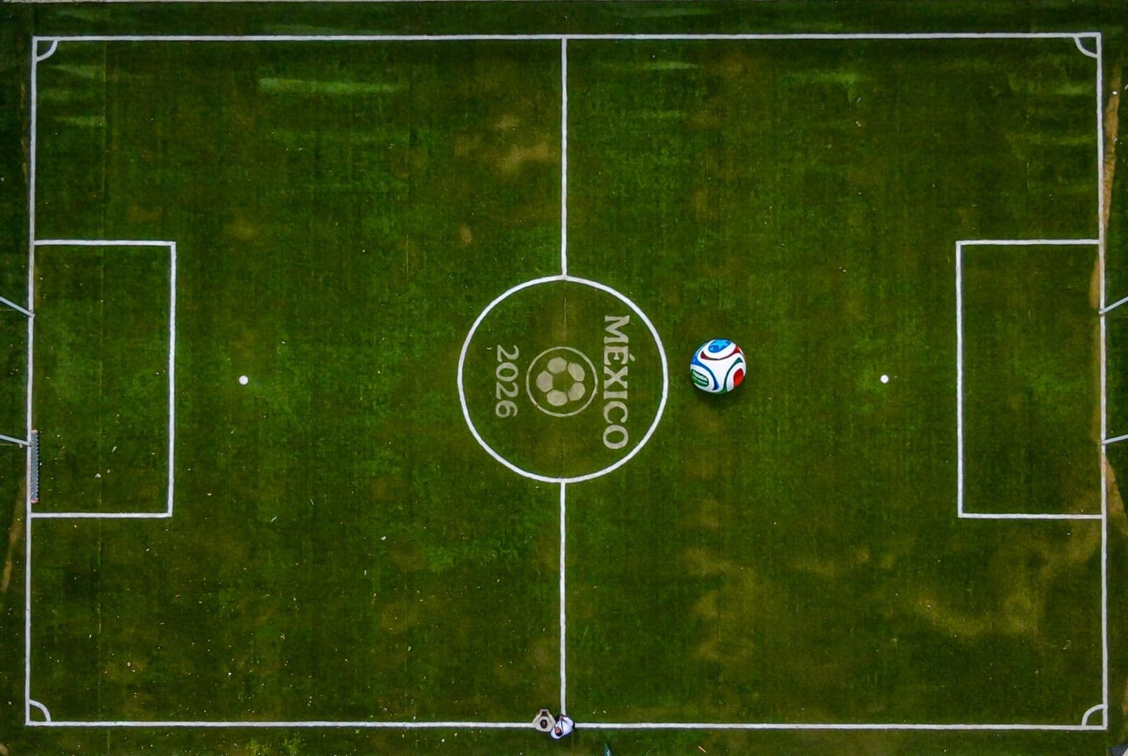Campo de fútbol con el logo de México 2026 en el centro y un balón en la zona de juego.