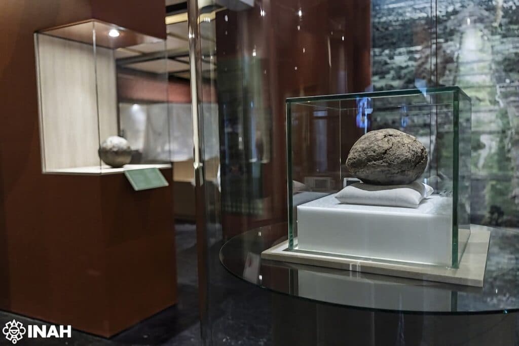Pelota de hule de 3,600 años exhibida en vitrina en la exposición Hule ritual en Teotihuacan, resaltando la cultura mesoamericana.