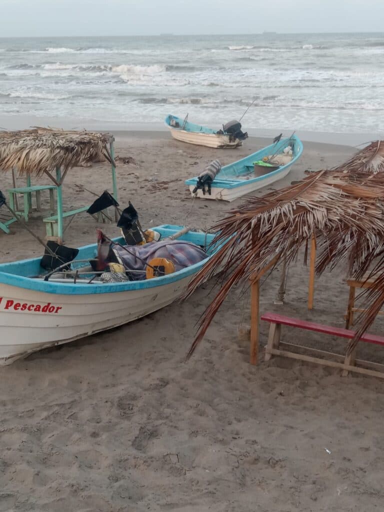 Botes de pesca varados en la arena de Playa Hermosa, con sombrillas de palma y olas del mar de fondo, reflejando la actividad turística local.