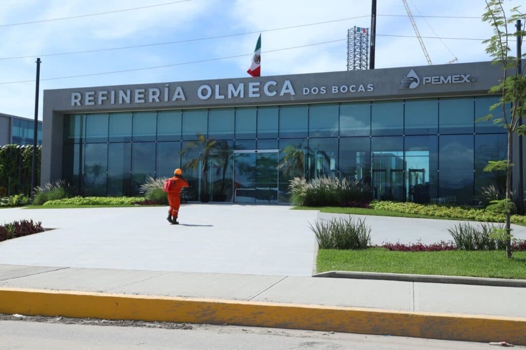 Trabajador ingresando a la Refinería Olmeca en Dos Bocas, Tabasco, con bandera mexicana ondeando y edificio de Pemex al fondo.