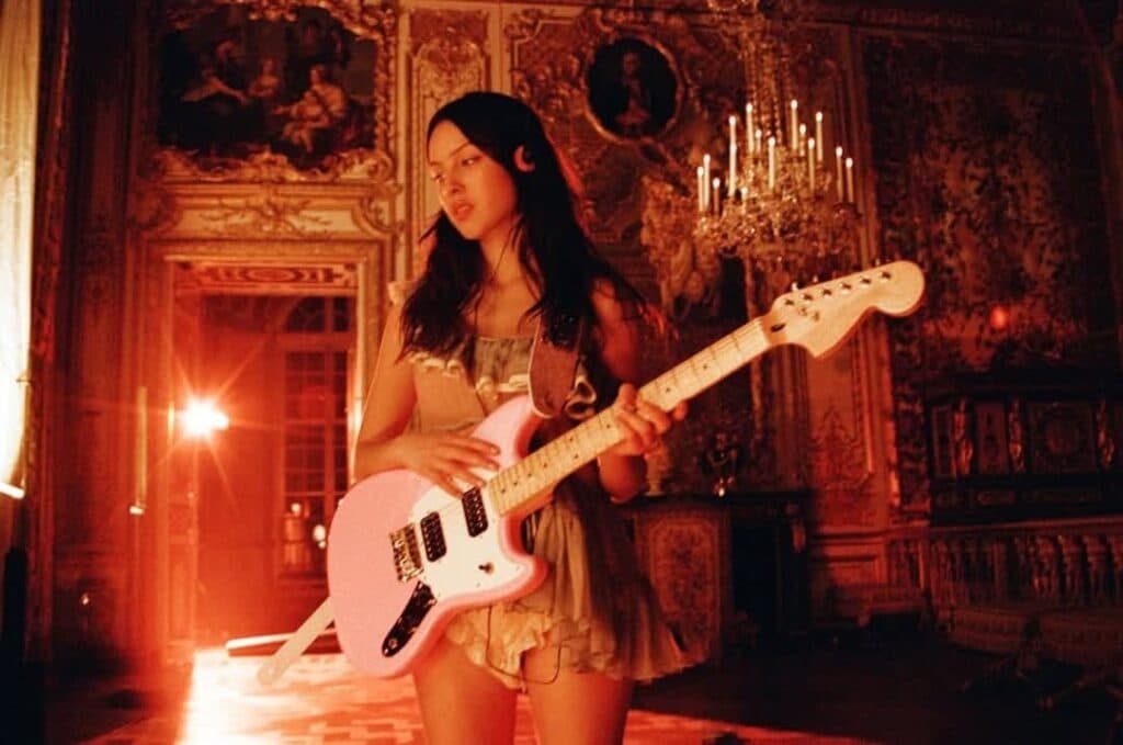 Mujer joven sostiene una guitarra rosa en un entorno elegante con candelabros, reflejando la esencia del festival benéfico de Olivia Rodrigo.