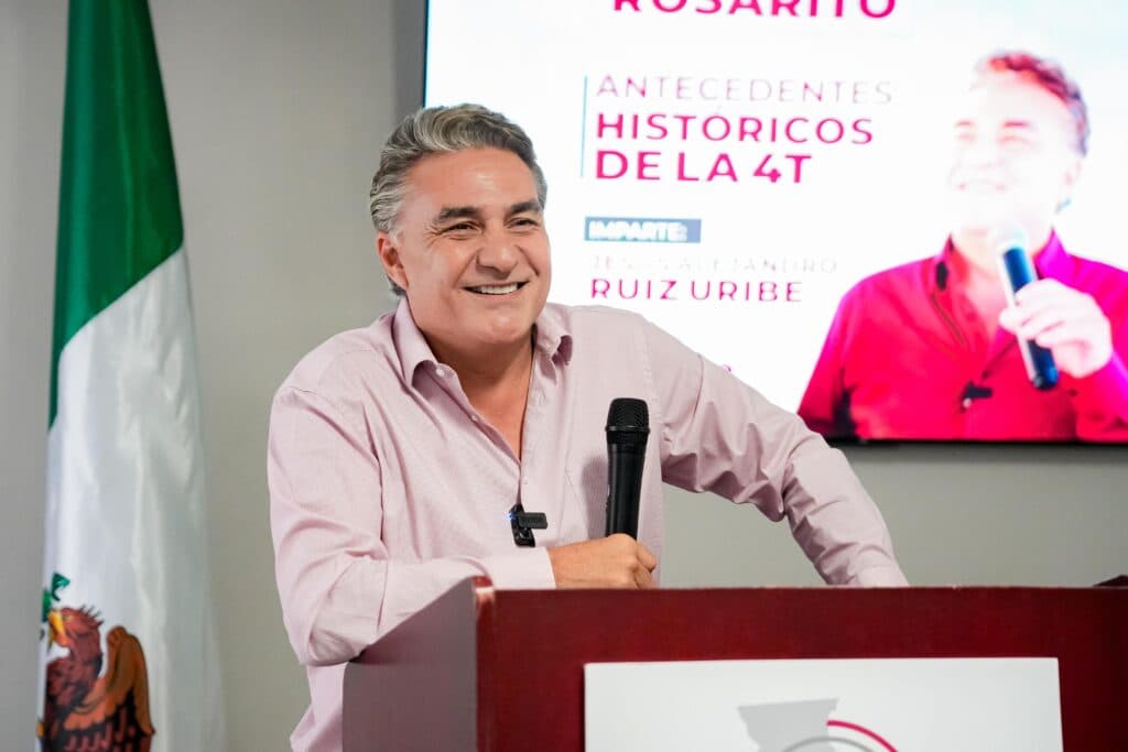 Jesus Ruiz Uribe sonríe mientras habla en un evento de MORENA en Rosarito, con una bandera mexicana de fondo y una