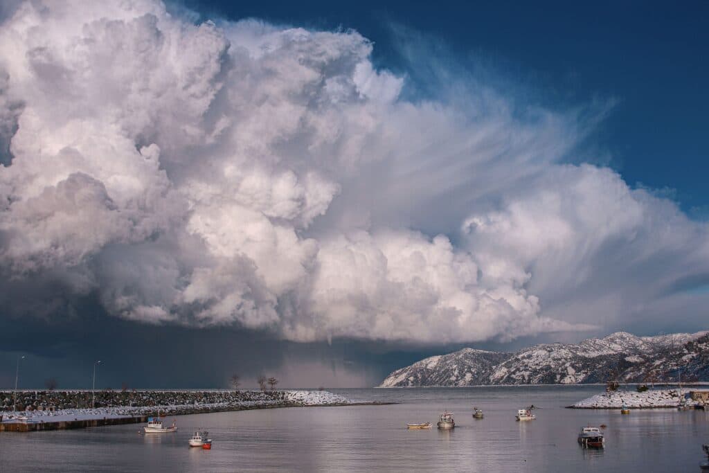 Nubes densas y amenazantes sobre un puerto, con barcos en el agua y montañas nevadas al fondo, reflejando cambios climáticos.