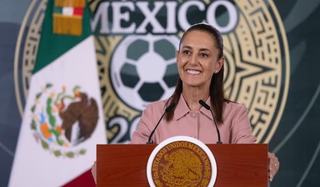 Claudia Sheinbaum Pardo sonríe mientras entrega boletos del Mundial 2026 a ciudadanos, con la bandera de México de fondo.