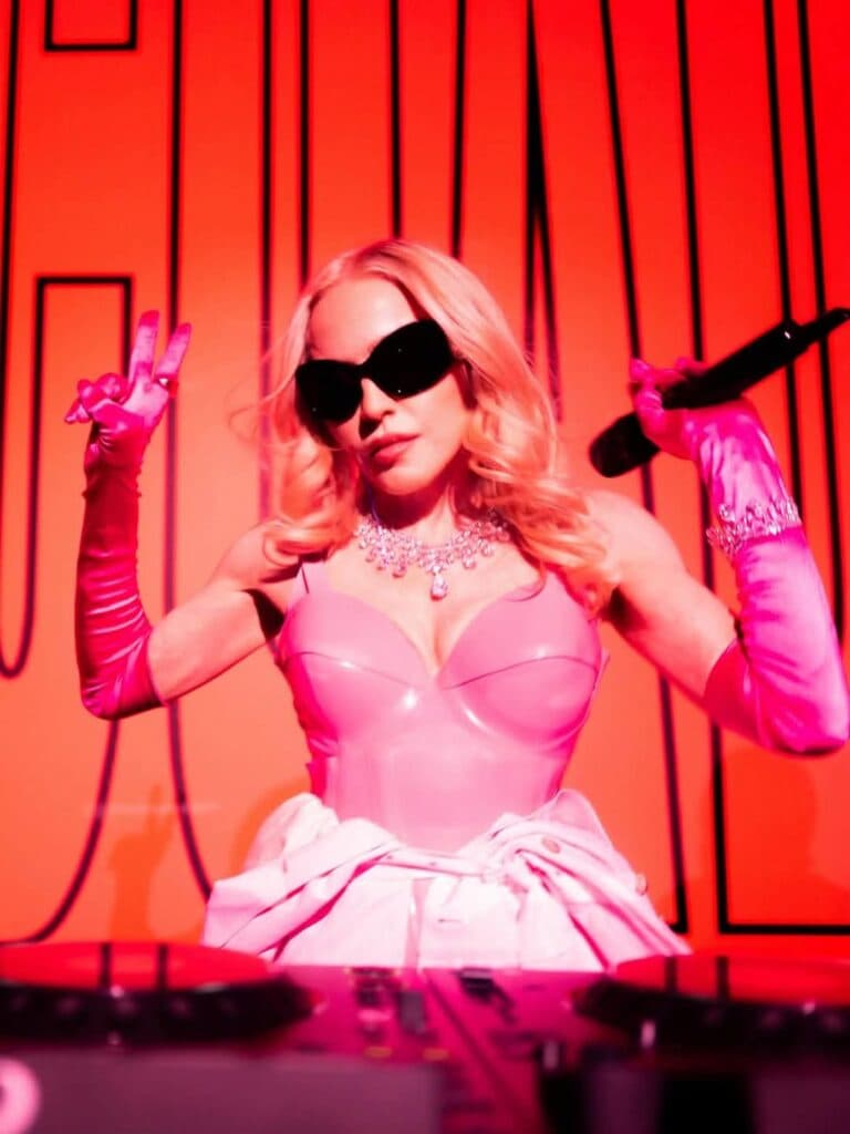 Madonna con atuendo rosa y gafas oscuras, sostiene un micrófono mientras actúa en un escenario iluminado en rojo.