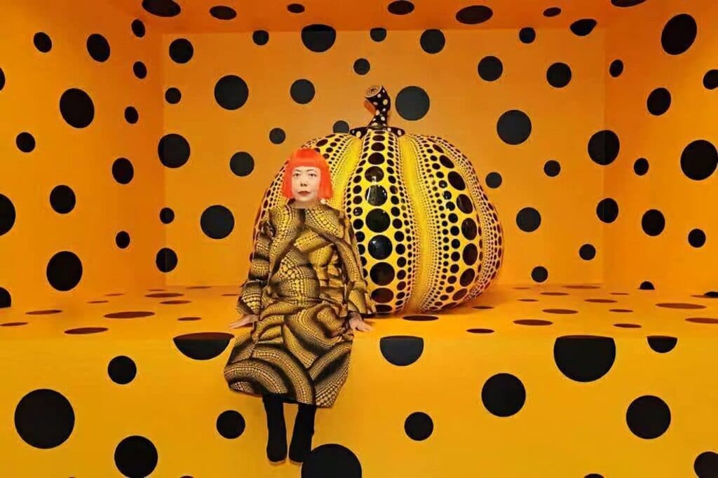 Yayoi Kusama sentada en una habitación amarilla con lunares negros, junto a una calabaza decorada con el mismo patrón, símbolo de su arte.