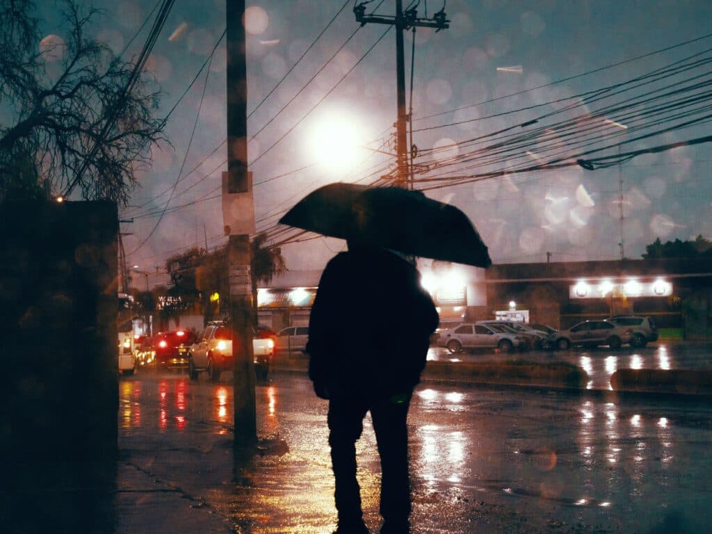 Persona con paraguas en la calle durante una tormenta eléctrica, con lluvia intensa y luces de vehículos reflejadas en el pavimento.