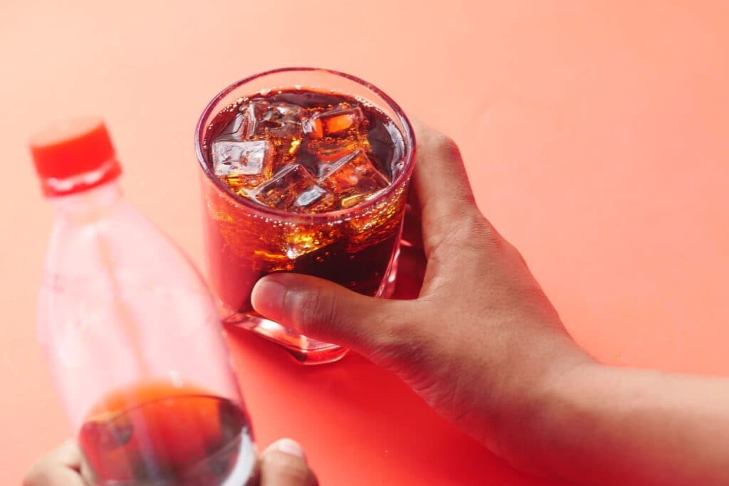 Vaso con refresco oscuro y hielo, mientras una mano sostiene una botella de bebida azucarada, resaltando el riesgo para la salud hepática.