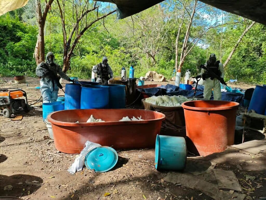 Autoridades en un laboratorio clandestino desmantelado en Michoacán, rodeados de barriles y equipos utilizados para la producción de drogas.