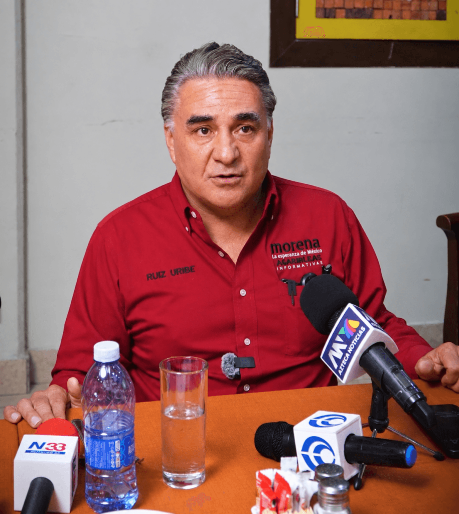 Ruiz Uribe durante conferencia de prensa, destaca logros de Claudia Sheinbaum en Baja California, con micrófonos y agua en la mesa.