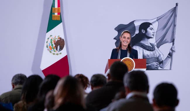 Claudia Sheinbaum en conferencia de prensa matutina en Palacio Nacional, con la bandera de México y un mural de una mujer sosteniendo la bandera.