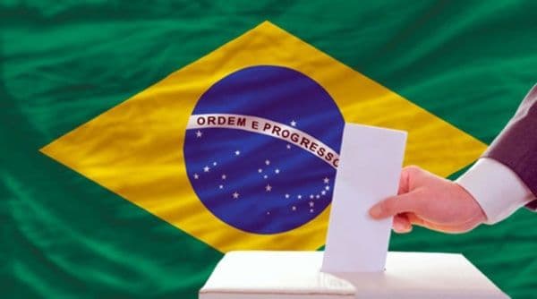 Una mano deposita un voto en una urna electoral frente a la bandera de Brasil, simbolizando la elección presidencial.