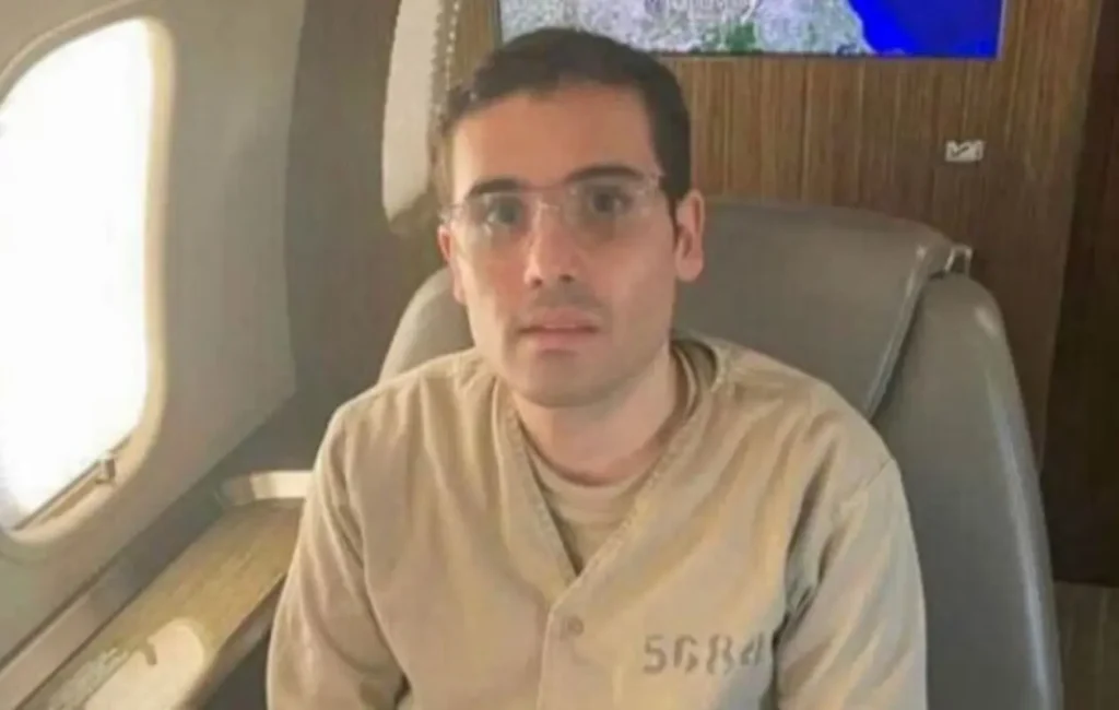 Ovidio Guzmán López, hijo de Joaquín "El Chapo" Guzmán, sentado en un avión privado, vestido con uniforme de prisionero.