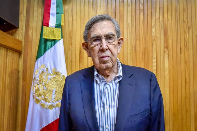 Cuauhtémoc Cárdenas, político y fundador del PRD, posando frente a la bandera de México en un ambiente institucional.